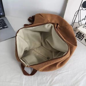 Puede incluir: Mochila de lona marr&oacute;n abierta con interior beige. La mochila tiene cierre de cremallera y correas marrones. El interior cuenta con un bolsillo con cremallera. La bolsa est&aacute; vac&iacute;a y lista para ser llenada.