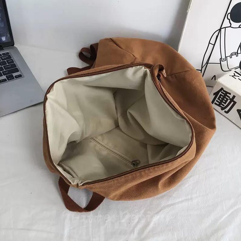 Puede incluir: Mochila de lona marr&oacute;n abierta con interior beige. La mochila tiene cierre de cremallera y correas marrones. El interior cuenta con un bolsillo con cremallera. La bolsa est&aacute; vac&iacute;a y lista para ser llenada.