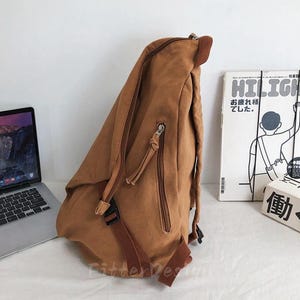 Puede incluir: Una mochila de lona marr&oacute;n con m&uacute;ltiples compartimentos con cremallera y correas ajustables. La mochila tiene un asa superior y un cierre de hebilla. Un port&aacute;til y un libro est&aacute;n en el fondo.