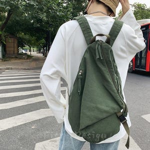 Puede incluir: Mochila de lona verde oliva con cremallera diagonal y correas ajustables. La mochila tiene un peque&ntilde;o bolsillo con cremallera en la parte delantera y un cierre de hebilla. La bolsa se lleva sobre el hombro.