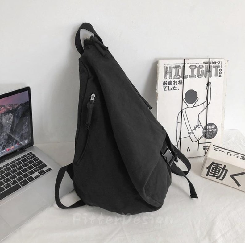 Puede incluir: Bandolera negra con una sola correa para el hombro y m&uacute;ltiples compartimentos con cremallera. La bolsa est&aacute; hecha de una tela duradera y tiene un cierre de hebilla. Un port&aacute;til y un libro con texto japon&eacute;s est&aacute;n en el fondo.