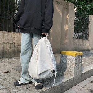 Puede incluir: Una mochila blanca con un bolsillo frontal con cremallera y un asa superior. La mochila tiene forma redondeada y cierre de cord&oacute;n. La persona lleva una sudadera con capucha negra y vaqueros azul claro.