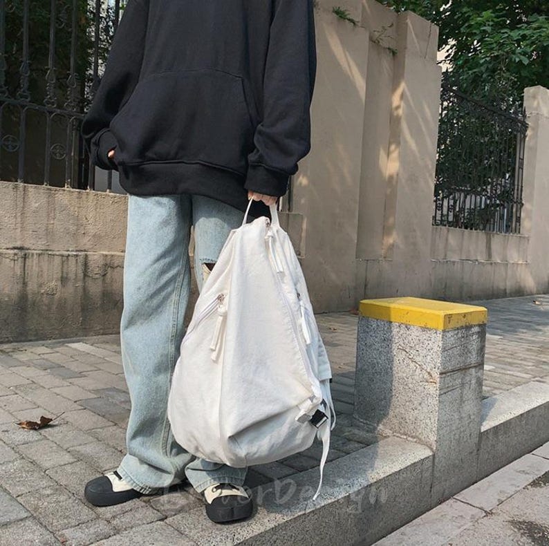 Puede incluir: Una mochila blanca con un bolsillo frontal con cremallera y un asa superior. La mochila tiene forma redondeada y cierre de cord&oacute;n. La persona lleva una sudadera con capucha negra y vaqueros azul claro.