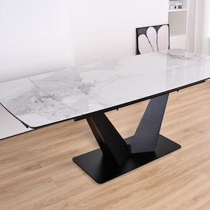 May include: A contemporary dining table featuring a white marble-effect surface and a black V-shaped base. The table has a rectangular shape with rounded corners. The table is approximately 183 cm long and 91 cm wide.