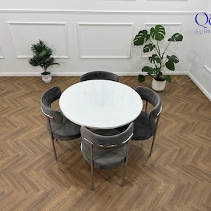 May include: A circular white marble-effect dining table with four grey velvet chairs with silver metal legs. The chairs are positioned around the table. The room features a wooden floor and white walls with decorative moulding. Plants are in the background.