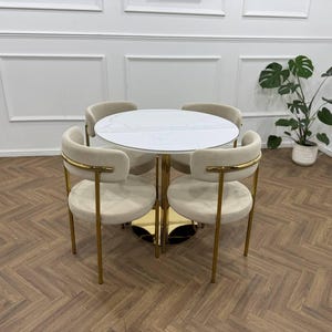 May include: A contemporary dining set with a round white marble-effect table and four cream-coloured chairs. The chairs have gold-coloured metal frames and a curved back design. The table has a gold-coloured base.
