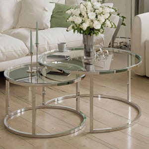 May include: Two circular coffee tables with mirrored glass tops and polished chrome frames. A vase of white flowers and a mug are on the larger table. The tables are nested, with the smaller one inside the larger one. The larger table has a diameter of approximately 90 cm.
