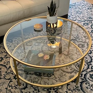 May include: A circular coffee table with a gold metal frame and two clear glass surfaces. A small potted plant, a coaster, and a book titled "Monet" are on the table. The table is 91 cm in diameter.