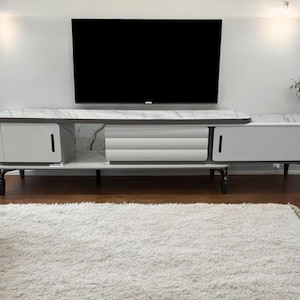 May include: A contemporary white and grey TV unit with a marble-effect top and black legs. The unit features two cabinets with vertical handles and a central ribbed section. A large black television is mounted above.