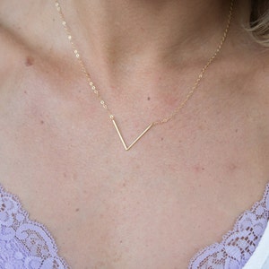 Könnte beinhalten: Eine goldene Halskette mit einem schlichten V-förmigen Anhänger. Die Kette wird von einer Person mit hellem Teint getragen und sitzt mittig auf der Brust.