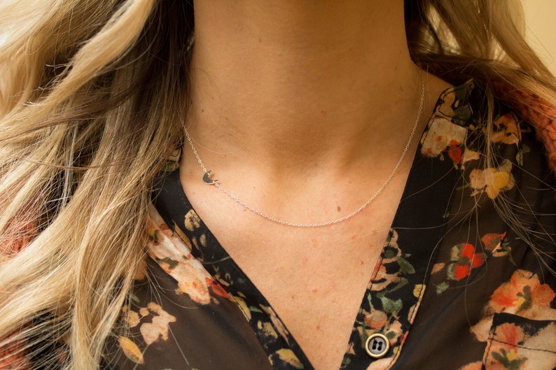 May include: A silver necklace with a crescent moon pendant. The necklace is worn by a person with long blonde hair and a floral patterned shirt.