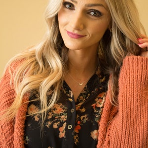 May include: A woman with long blonde hair is wearing a coral cardigan over a black floral blouse. She is wearing a delicate silver necklace with a crescent moon pendant.