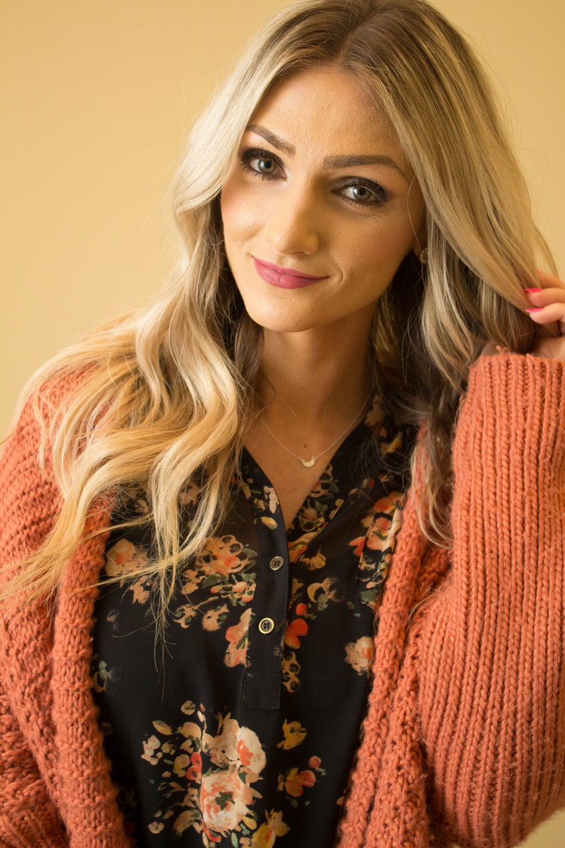 May include: A woman with long blonde hair is wearing a coral cardigan over a black floral blouse. She is wearing a delicate silver necklace with a crescent moon pendant.