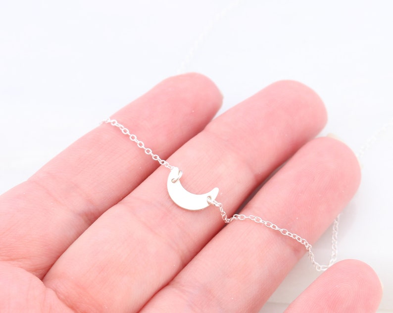 May include: A silver crescent moon charm necklace on a delicate silver chain. The necklace is displayed on a hand.