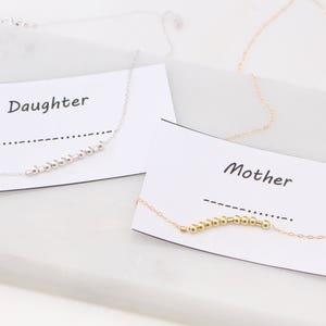May include: Two necklaces with cards that say "Daughter" and "Mother". The necklace on the left has silver beads and the necklace on the right has gold beads.