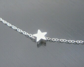 Puede incluir: Un delicado collar de plata con un pequeño colgante de estrella tridimensional. La estrella está centrada en una fina cadena, creando un diseño minimalista y elegante. El collar se muestra sobre un fondo oscuro.
