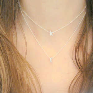 May include: Two delicate silver necklaces layered on a person's neck. The top necklace features the letter "k", and the bottom necklace has the letter "f". The necklaces are simple and elegant, suitable for everyday wear.