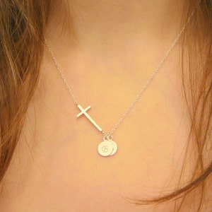 Puede incluir: Un collar de plata con un colgante de cruz lateral y dos pequeños abalorios redondos. Los abalorios tienen una letra 'B' grabada.