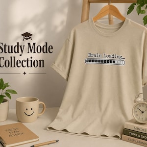 Op de afbeelding: Een beige t-shirt met korte mouwen voorzien van de zwarte tekst Brain Loading... boven een laadbalk-afbeelding. Het shirt wordt getoond op een houten kledinghanger, met de tekst Study Mode Collection zichtbaar aan de linkerzijde.