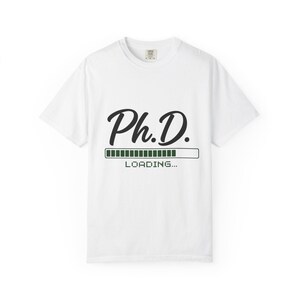May include: White t-shirt with the text "Ph.D. LOADING..." in black script and a green loading bar graphic. The shirt is a crew neck style and appears to be made of a soft material.