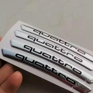Könnte beinhalten: Vier silberne Quattro-Autoembleme mit schwarzer Schrift. Die Embleme sind gebogen und haben ein metallisches Finish. Das Wort "Quattro" ist auf jedem Emblem deutlich sichtbar.