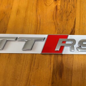 Peut inclure: Un emblème TT RS argenté avec un accent rouge, monté sur une base rectangulaire blanche. L'emblème est conçu pour une utilisation automobile, probablement pour l'extérieur d'une voiture. Les lettres sont métalliques et en trois dimensions.
