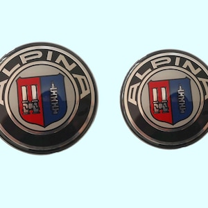 Peut inclure: Deux emblèmes circulaires Alpina. Chaque emblème présente un centre argenté avec un motif de bouclier rouge et bleu, entouré d'un anneau noir avec le mot "ALPINA" en argent. Les emblèmes sont probablement destinés à l'automobile.