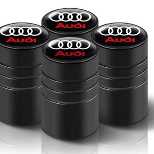 Könnte beinhalten: Vier schwarze Reifenventilkappen mit dem Audi-Logo in Weiß und dem Audi-Namen in Rot. Die Kappen haben eine glänzende Oberfläche und eine zylindrische Form, für den Automobilbereich.