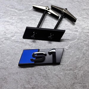 Peut inclure: Un emblème S1 noir et bleu avec une finition métallique. L'emblème comprend deux boulons de montage en métal filetés avec des attaches à ailettes de type papillon fixées à l'arrière pour une installation sécurisée sur une calandre.
