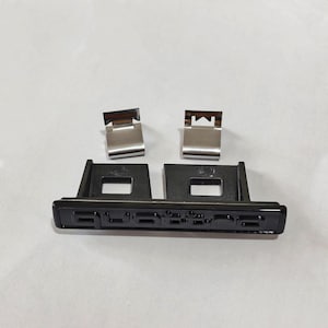 May include: Black plastic car emblem with the word "QUATTRO" in silver lettering. Two silver metal clips are above the emblem. The emblem is rectangular with a glossy finish.