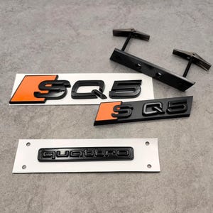 Peut inclure: Emblèmes de voiture SQ5 et Quattro noirs et orange. Les emblèmes SQ5 ont une finition noire avec des accents orange. L'emblème Quattro est noir. Les emblèmes sont sur un support blanc.