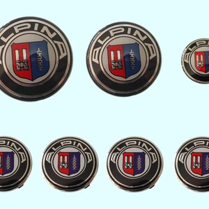 Peut inclure: Plusieurs enjoliveurs de roue Alpina. Les enjoliveurs sont ronds avec un bord noir, un centre argenté et le logo Alpina au centre. Le logo présente un écusson rouge et bleu avec le mot "ALPINA" en argent.