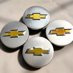 May include: Four silver-colored hubcaps with a gold and silver bowtie logo in the center. The hubcaps are round and have a metallic finish. The logo is a prominent feature, with a bright yellow center and silver outline.