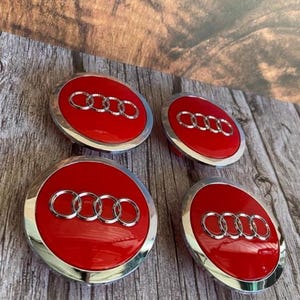 Peut inclure: Quatre enjoliveurs de roue de voiture rouges et argentés avec le logo Audi. Chaque capuchon présente un centre rouge brillant avec le logo des anneaux Audi argentés et un bord extérieur argenté. Les capuchons sont disposés sur une surface en bois.
