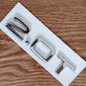 Peut inclure: Emblème 2.0T chromé sur un support blanc, posé sur un fond imitation bois. Les lettres métalliques reflètent la lumière. Le design comprend un '2', un petit carré, un 'O' et un 'T'.