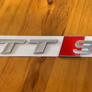 Peut inclure: Un emblème TT S chromé, avec les lettres "TT" et le chiffre "S", est présenté sur un support rectangulaire blanc. Le "S" est placé sur un fond rouge. L'emblème est conçu pour une utilisation automobile.