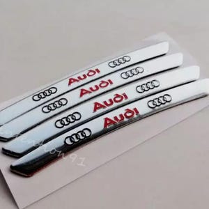 Könnte beinhalten: Vier silberfarbene Audi-Autoembleme mit dem Audi-Logo und dem Wort "Audi" in Rot. Die Embleme sind rechteckig und leicht gebogen, mit glänzendem Finish. Sie liegen auf einem weißen Hintergrund.