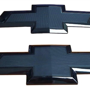 May include: Two black Chevrolet bow tie emblems. The top emblem is smaller than the bottom one. Both emblems have a glossy black finish with a textured center and a chrome-like border. The background is white.