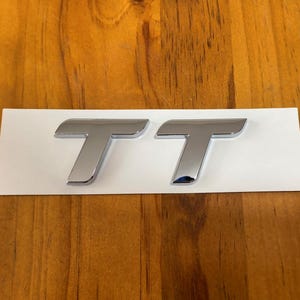 Peut inclure: Deux emblèmes "TT" chromés, probablement pour un véhicule, sont présentés sur un support rectangulaire blanc. Les emblèmes ont un design élégant et moderne avec une surface réfléchissante, sur un fond en bois chaud.