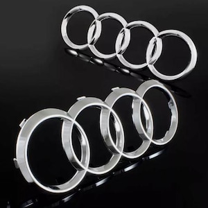 Peut inclure: Deux logos chromés Audi, chacun composé de quatre anneaux entrelacés. Les emblèmes sont présentés sur un fond noir, l'un au-dessus de l'autre. Les anneaux ont une finition métallique réfléchissante, créant un aspect élégant.
