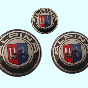 Peut inclure: Trois badges Alpina. Les badges circulaires ont un anneau extérieur noir, un anneau intérieur argenté et un emblème central avec des sections rouges et bleues. Le mot "ALPINA" est en lettres argentées.