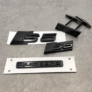 Könnte beinhalten: Schwarze S5- und Quattro-Autoembleme und ein Paar schwarze Manschettenknöpfe. Die S5-Embleme sind in glänzendem Schwarz gehalten, das Quattro-Emblem ebenfalls in Schwarz. Die Manschettenknöpfe haben eine rechteckige schwarze Basis und silberne Akzente.