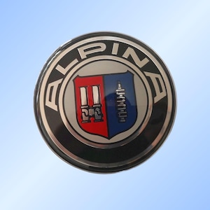 Peut inclure: Un emblème rond argenté et noir avec le mot "ALPINA" en lettres argentées sur le bord extérieur. L'emblème présente un écusson avec des sections rouges et bleues, contenant des illustrations mécaniques détaillées.