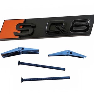 May include: Black and orange S Q8 emblem with mounting hardware. The emblem has a black background with the letters "S Q8" in a glossy finish. Includes two blue screws and two blue toggle bolts.