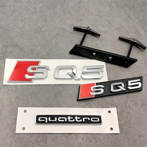 Peut inclure: Plusieurs emblèmes de voiture, dont des badges "SQ5" et "quattro". Les badges "SQ5" présentent une finition argentée avec des détails rouges, tandis que le badge "quattro" est noir avec des lettres argentées. Un support de montage noir est également présent.