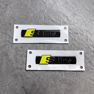 Peut inclure: Deux emblèmes rectangulaires S-line avec une finition noire brillante et un carré jaune vif sur le côté gauche. Chaque emblème est fixé sur un support en papier blanc rectangulaire doté de quatre trous circulaires aux coins pour la fixation.