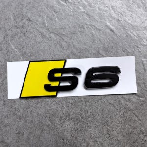 Peut inclure: Un emblème rectangulaire présentant le texte S6 en lettres noires brillantes. La lettre S est partiellement superposée sur une forme de parallélogramme jaune vif. L'ensemble est monté sur un support rectangulaire blanc à la finition nette.