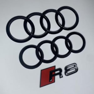 Peut inclure: Ensemble d'emblèmes Audi noirs, comprenant les quatre anneaux entrelacés et un badge R8 avec un fond carré rouge. Les emblèmes sont brillants et probablement en plastique ou en métal, pour une utilisation automobile.