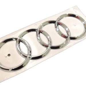 Peut inclure: Emblème chromé représentant les quatre anneaux entrelacés, symbole de la marque automobile Audi. L'emblème est présenté sur un support blanc, prêt à être appliqué. Les anneaux ont une finition polie et réfléchissante.
