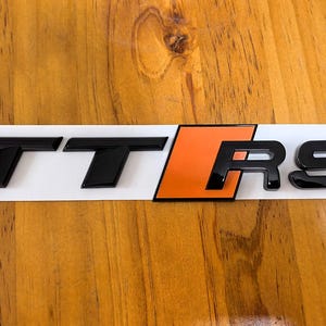 Peut inclure: Emblème noir TT RS avec un accent orange, sur fond blanc. L'emblème brillant est probablement un badge de voiture, conçu pour l'extérieur d'un véhicule.